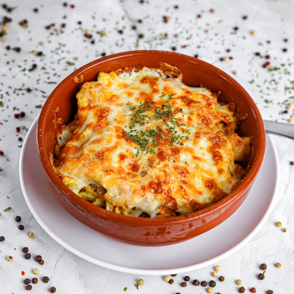 A baked dish with melted, bubbly cheese and green herbs in a rustic brown bowl.