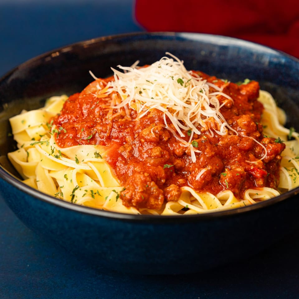Close-up of tagliatelle pasta with rich red bolognese sauce, topped with shredded cheese and parsley.