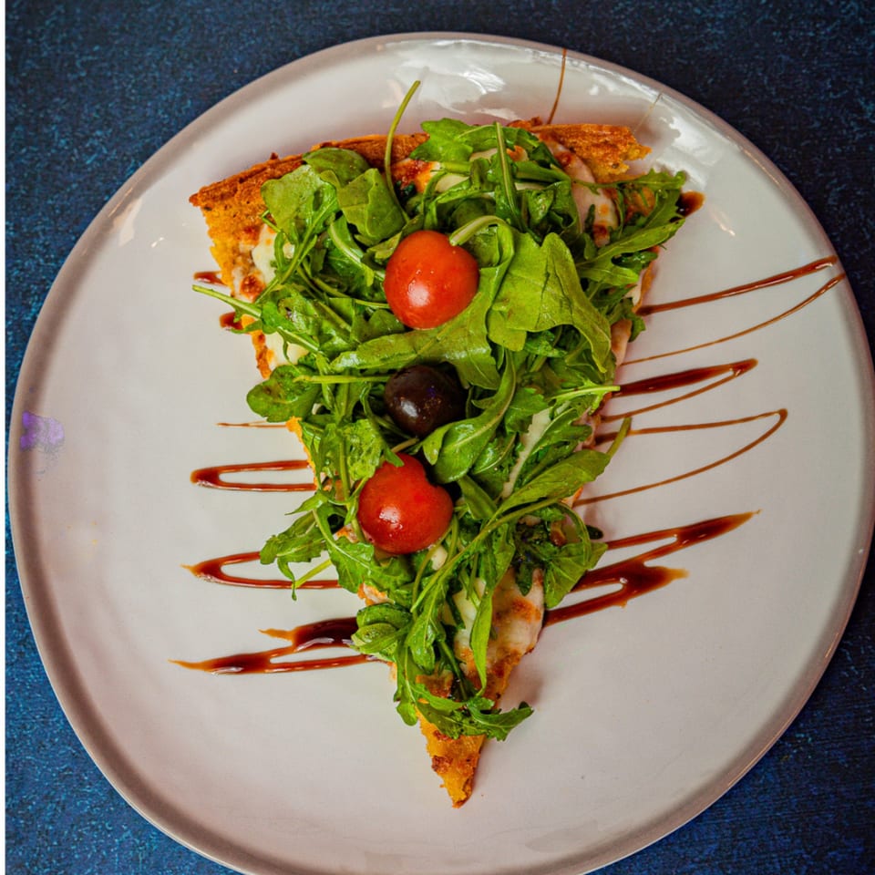 A slice of Argentinian pizza topped with fresh arugula, cherry tomatoes, and an olive.
