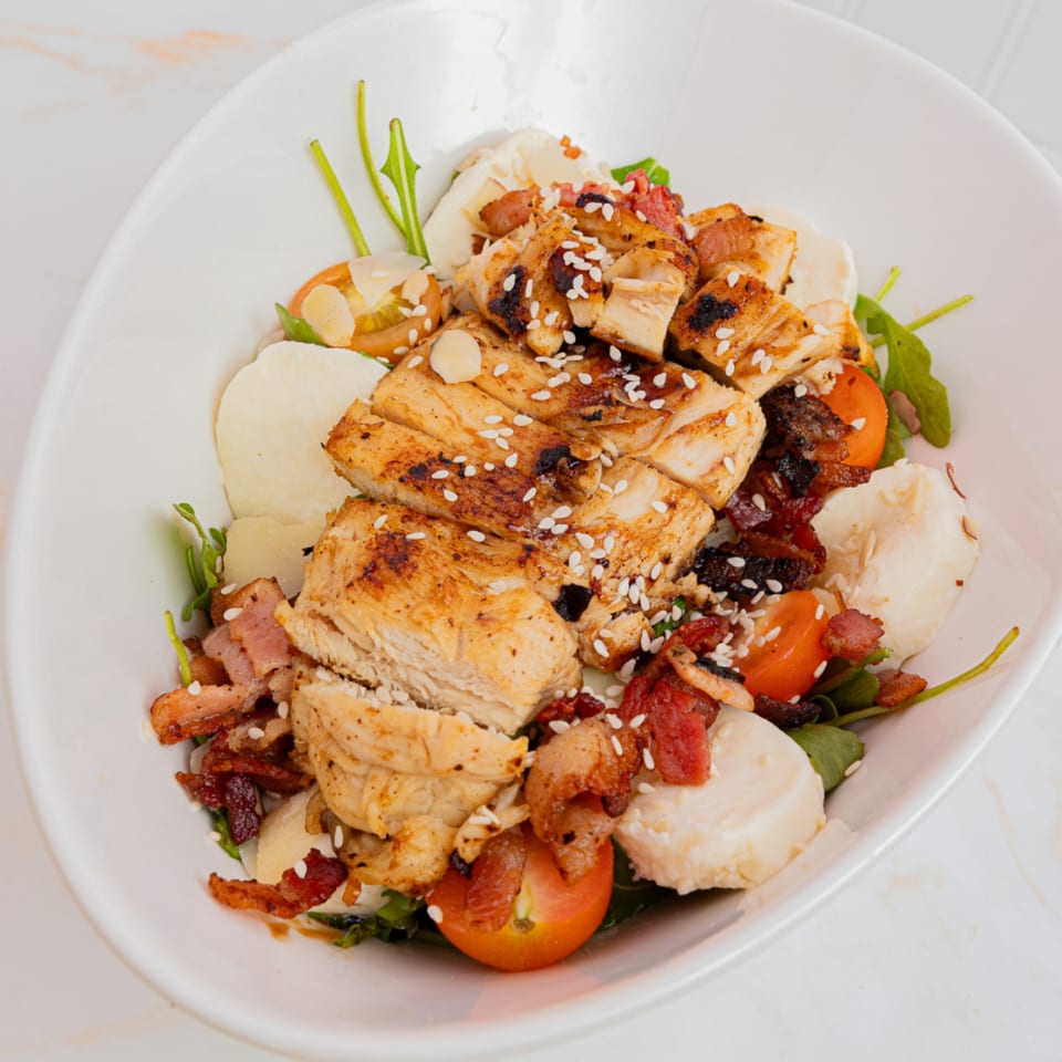 Grilled chicken salad with bacon, cherry tomatoes, mozzarella, and sesame seeds in a white bowl.
