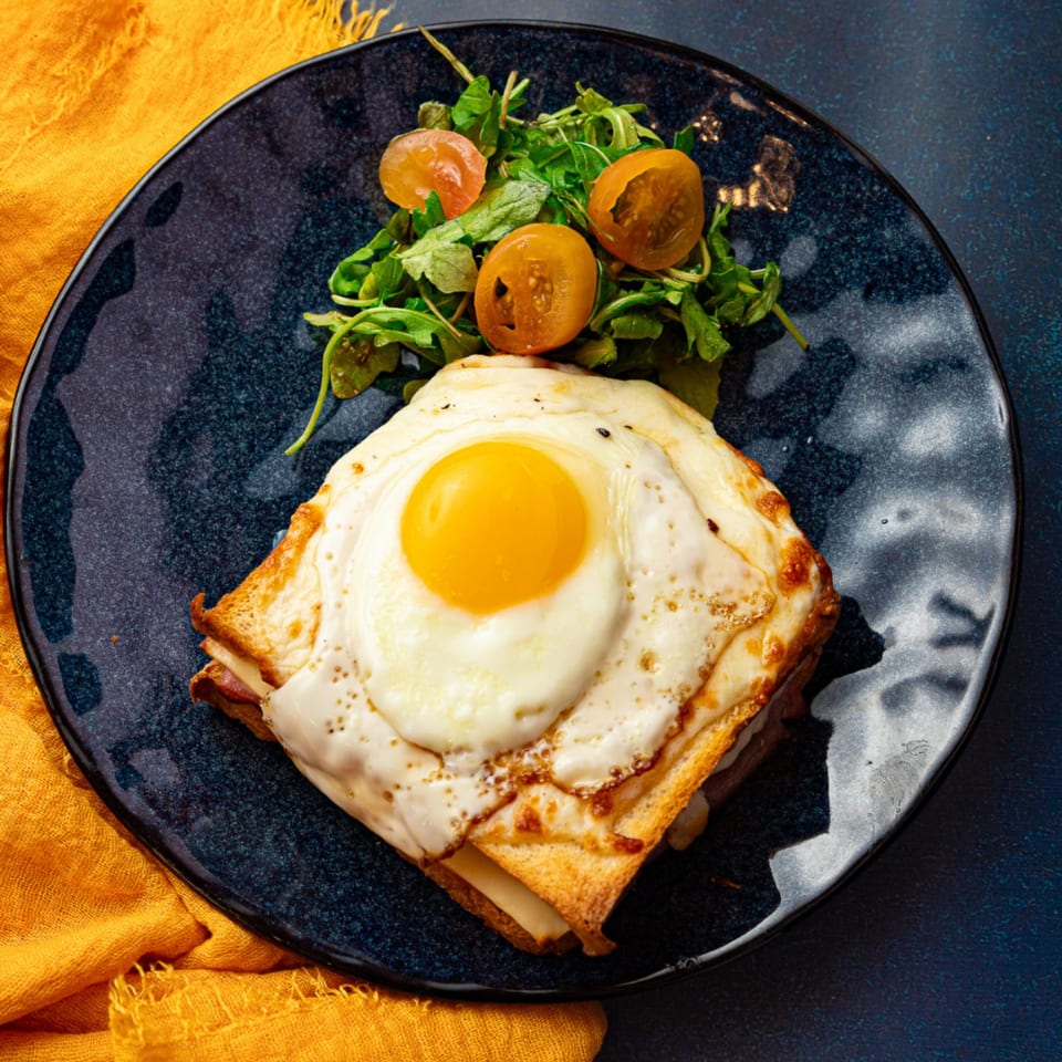 Vita Nostra Bakery Bistro's Croque Madame with a fried egg and side salad on a dark plate.